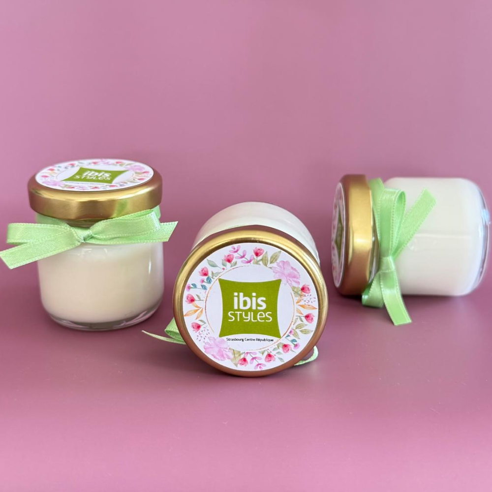 Bougies personnalisées avec logo Ibis Styles, pot en verre, couvercle doré et ruban vert pour cadeaux d'entreprise.